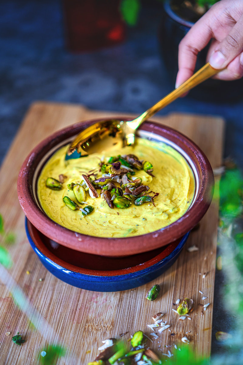 Close-up of shrikhand in a clay bowl with a golden spoon, garnished with nuts and a vibrant, colorful background.