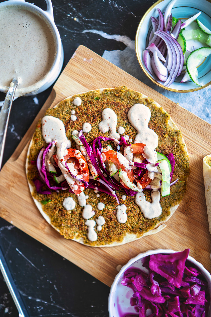 Top view of a smashed falafel wrap being assembled on a wooden board with toppings including red cabbage, onions, tomatoes, and cucumber, drizzled with tahini sauce.