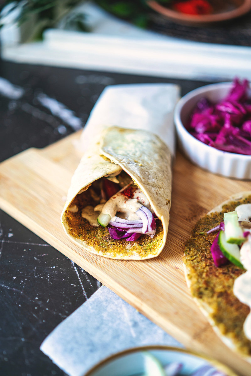 A close-up of a smashed falafel wrap filled with fresh vegetables and drizzled with a creamy sauce on a wooden board, with a side of purple cabbage.