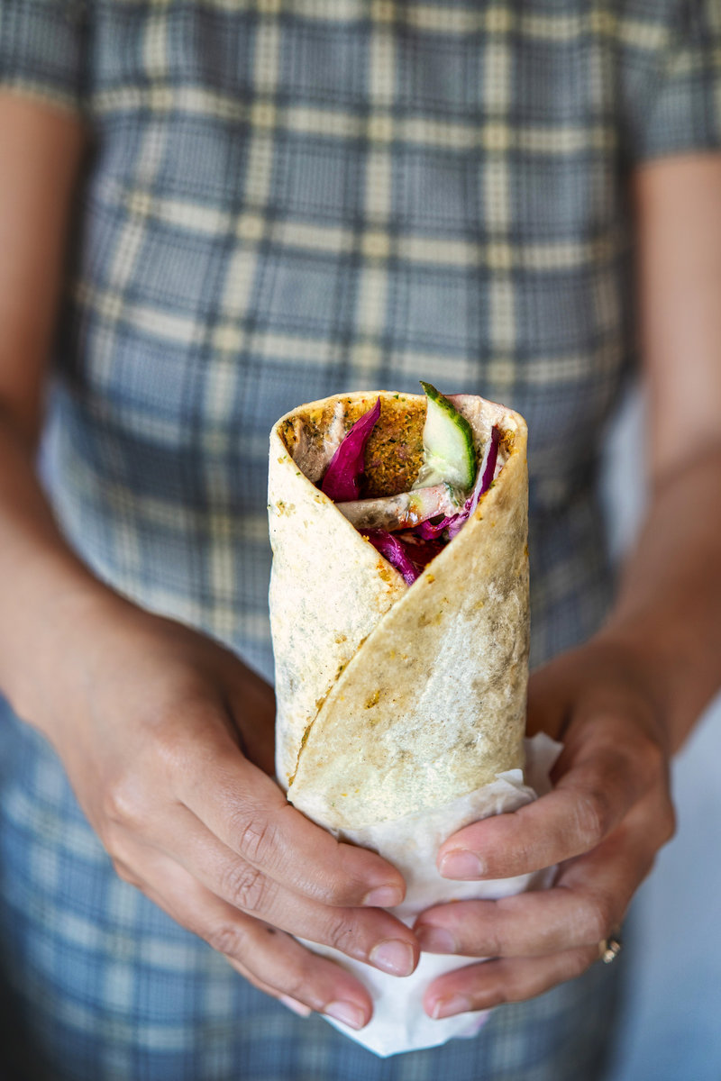 A person holding a wrapped Smashed Falafel Wrap, filled with vibrant vegetables and falafel, with a plaid background.