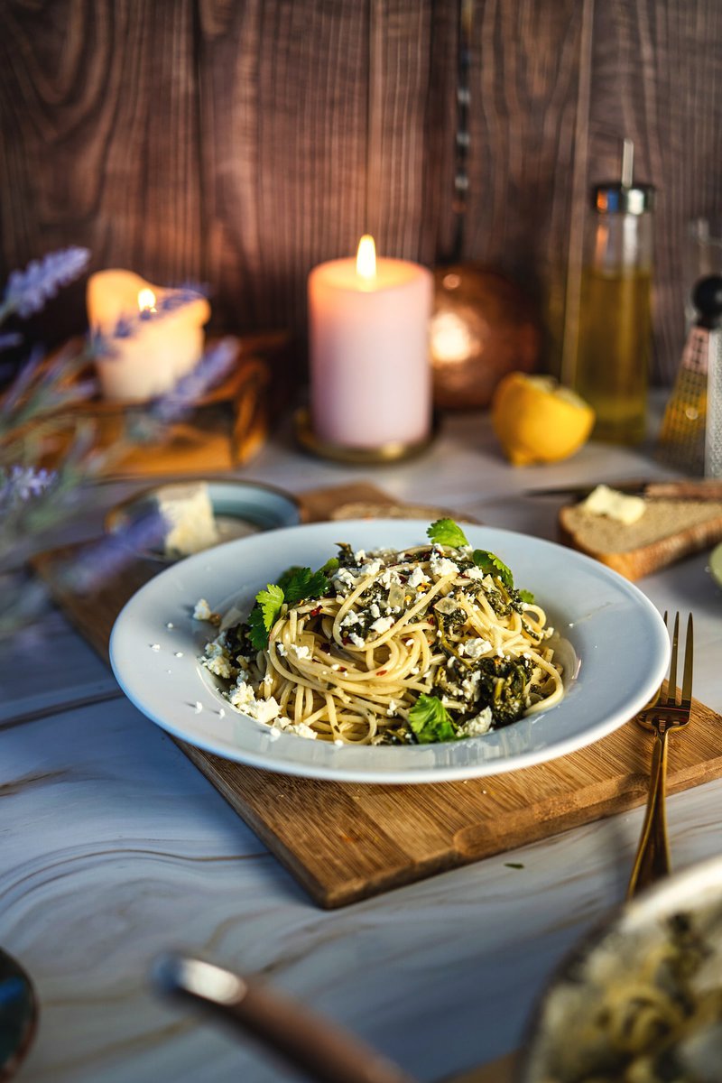 Spinach pasta served on light blue plate, garnished with parmesan cheese and herbs, placed on wooden board with ambient candle lighting and table decor