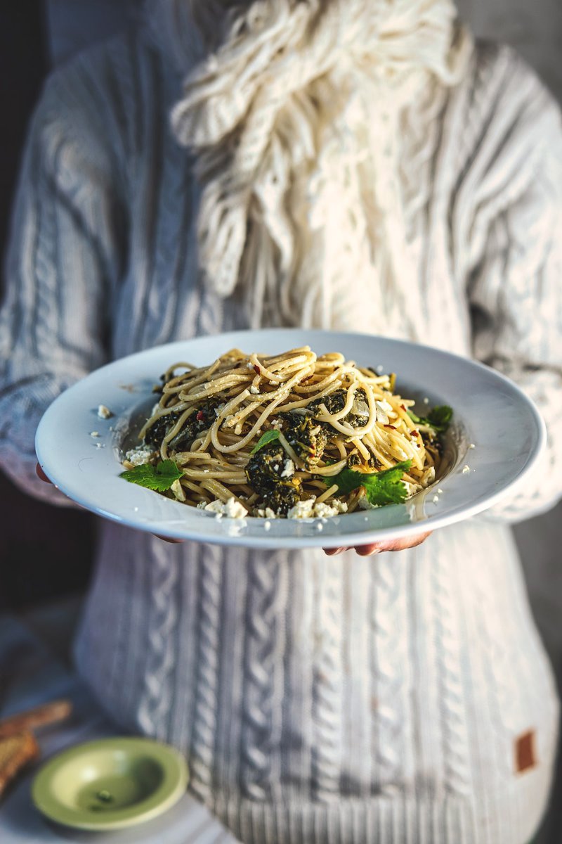 Person holding a white plate of spinach pasta, wearing a cozy sweater in a warm setting