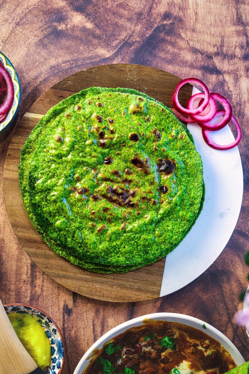 Stack of soft spinach rotis served on a wooden board with pickled onions and curry on the side