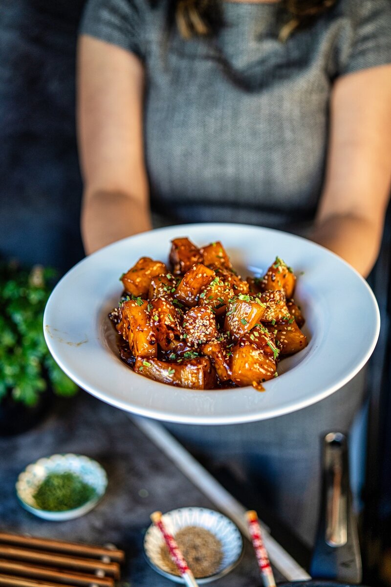 Hand holding a bowl of sticky chili garlic pineapple with glossy sauce, caramelized pineapple chunks, and sesame topping