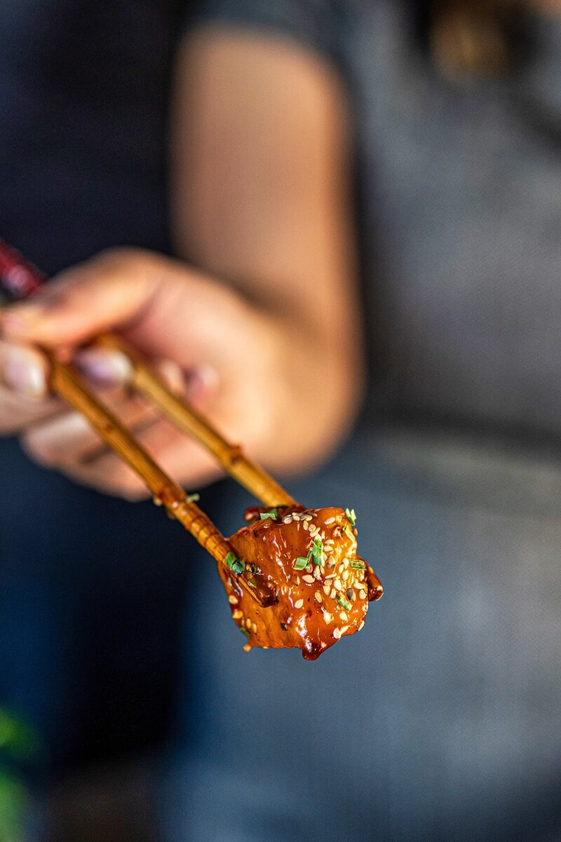 Close-up of sticky chili garlic pineapple held with chopsticks, showing juicy, glossy, caramelized pineapple in chili garlic sauce