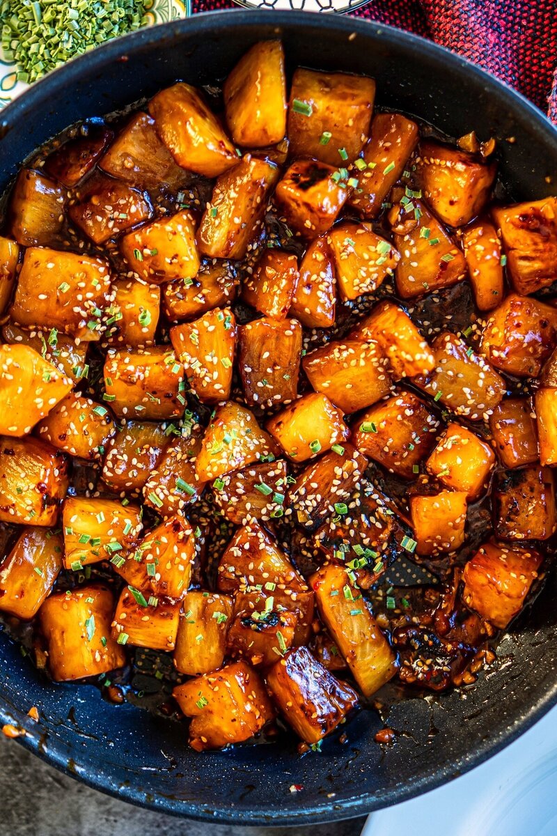 Sticky chili garlic pineapple cooking in a pan with glossy chili garlic sauce, caramelized edges, sesame seeds, and chives