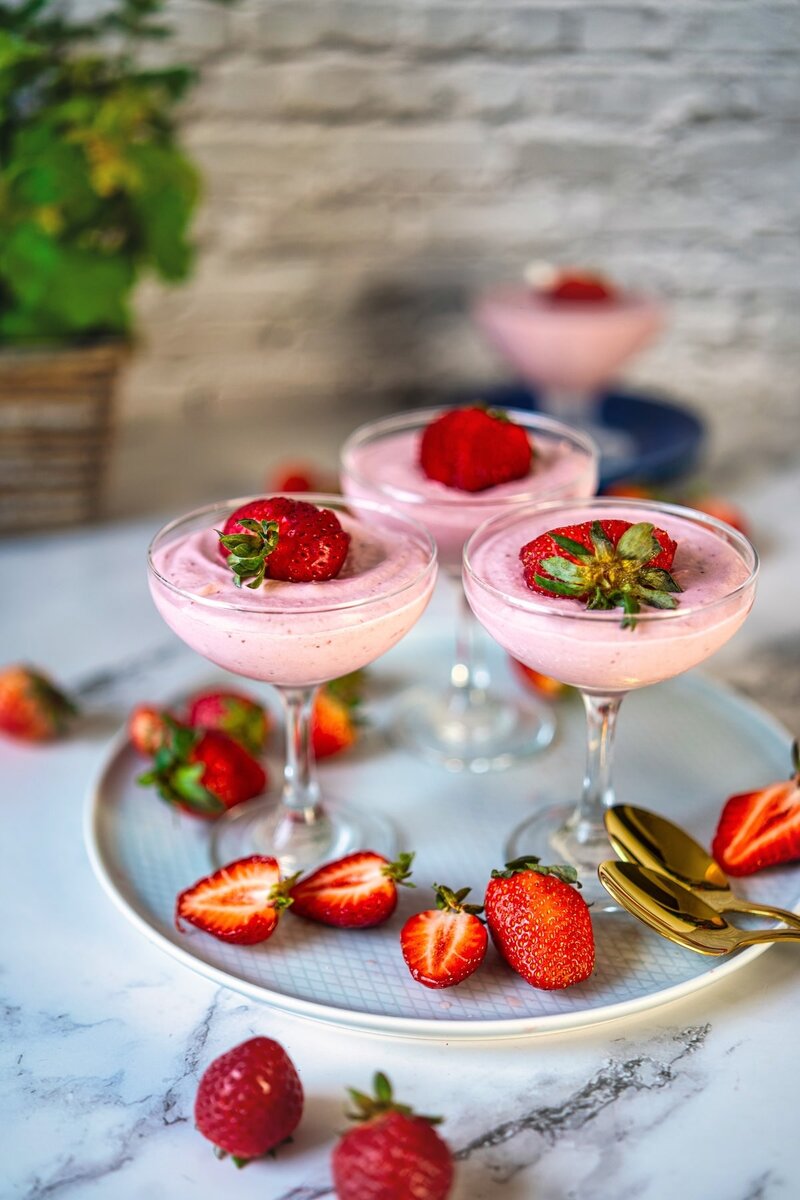 Glasses filled with strawberry mousse, topped with fresh strawberries, served on a tray with scattered strawberries on a marble surface.