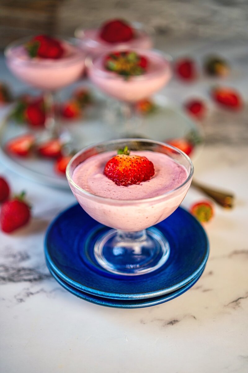 lose-up of a strawberry dessert in a coupe glass on a blue plate. The creamy pink mousse is topped with a whole strawberry, with additional dessert glasses and fresh strawberries visible in the background.