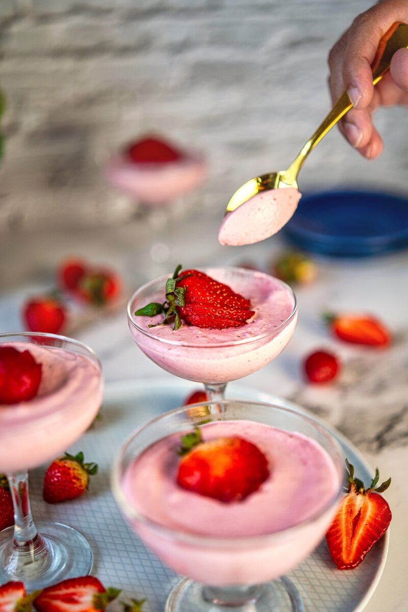 A gold spoon scooping creamy strawberry dessert from one of several coupe glasses arranged on a tray. Each glass contains pink mousse topped with fresh strawberries, creating an elegant dessert presentation.
