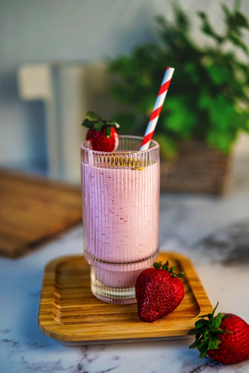 Tall glass of strawberry banana smoothie with red striped straw and fresh berries on wooden coaster