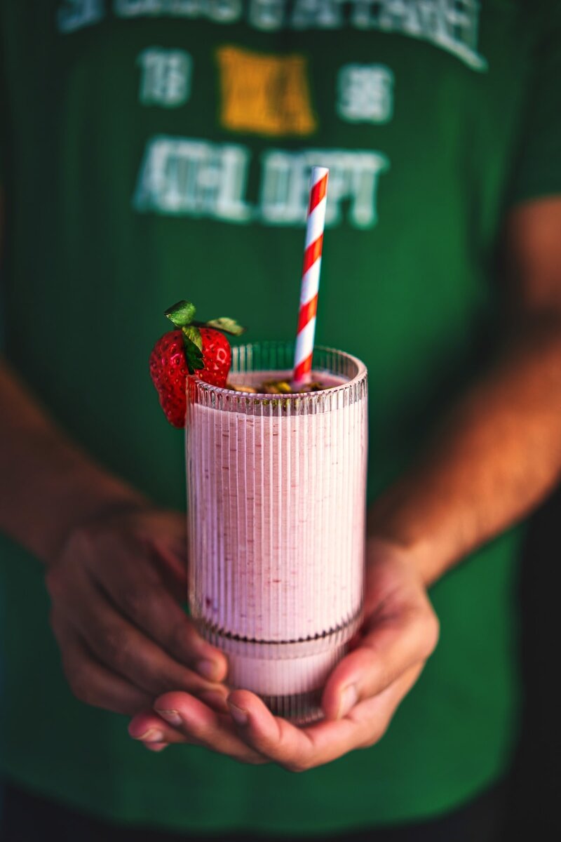 Hand holding a glass of homemade strawberry smoothie with fresh strawberries and striped straw – easy healthy breakfast recipe