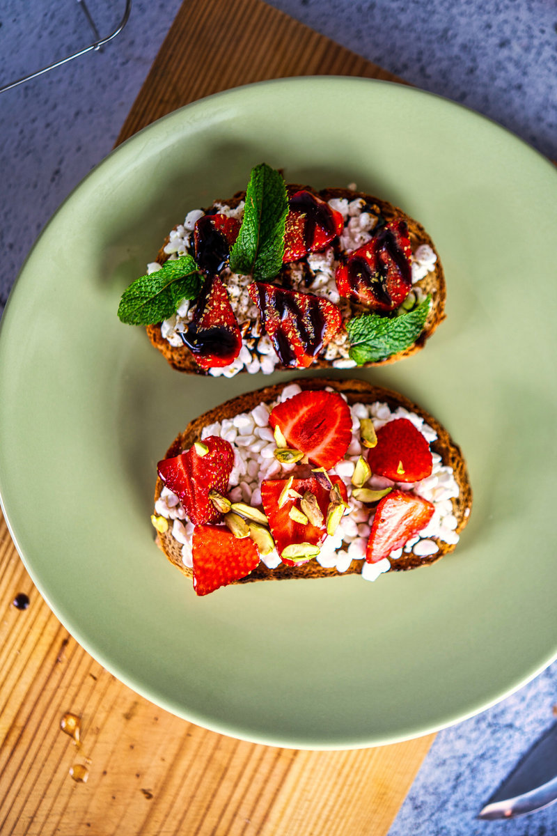 Overhead shot of two pieces of strawberry cottage cheese toast on a green plate; one with mint leaves and balsamic reduction, the other with fresh strawberries and pistachios.