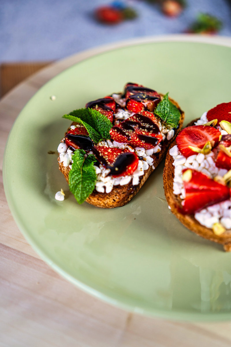 Side view of two pieces of strawberry cottage cheese toast on a green plate, one topped with fresh mint leaves and balsamic reduction, and the other with strawberries and pistachios.