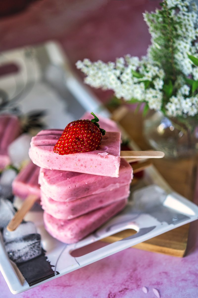 Strawberry creamsicles displayed on a white tray with fresh strawberries and a decorative flower arrangement in the background.