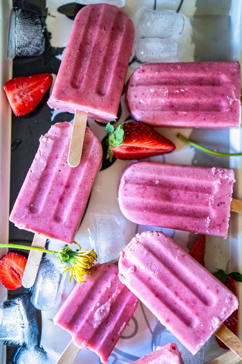 Assorted strawberry creamsicles surrounded by ice cubes and fresh strawberries on a reflective surface.