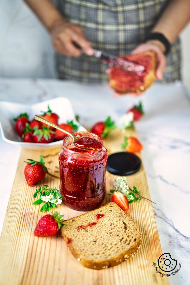 open jar of strawberry jam, with a golden spoon sticking out of it, with a female spreading jam on the bread in the background