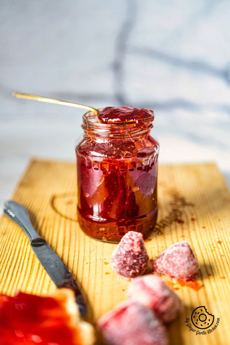 strawberry jam in a glass jar with with a golden spoon showing texture, and some frozen strawberries on the side