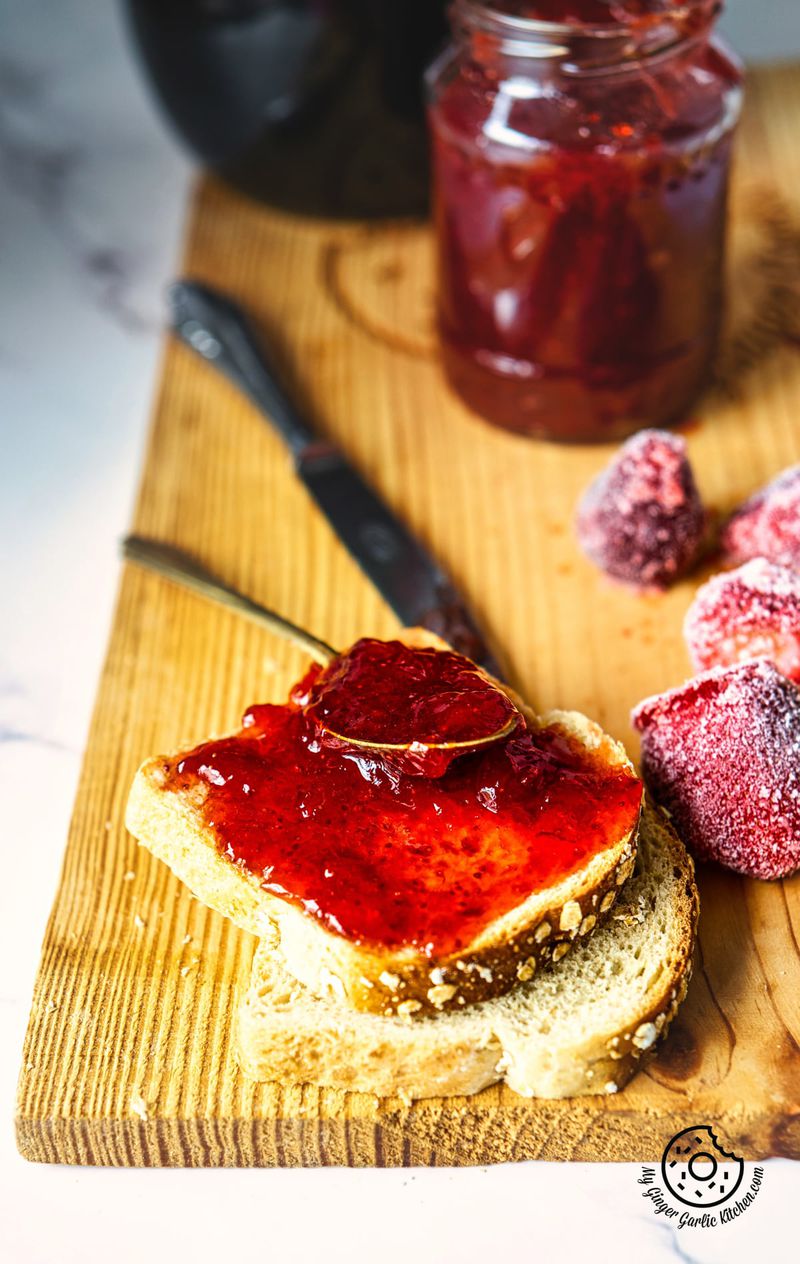 bread slices ith some strawberry jam on top, sitting on a wooden board, with an open jar of jam on the background