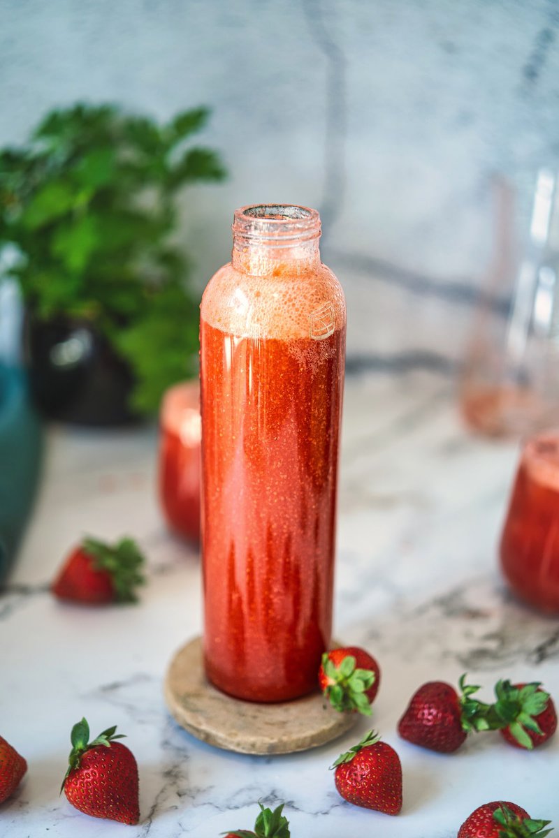 Glass bottle filled with vibrant red strawberry juice surrounded by fresh strawberries on a marble surface.