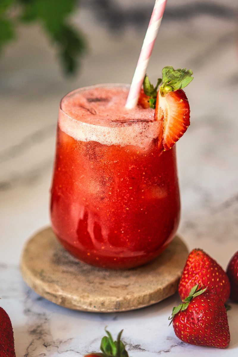 Close-up of a glass of strawberry juice with ice, garnished with a strawberry slice and a straw.