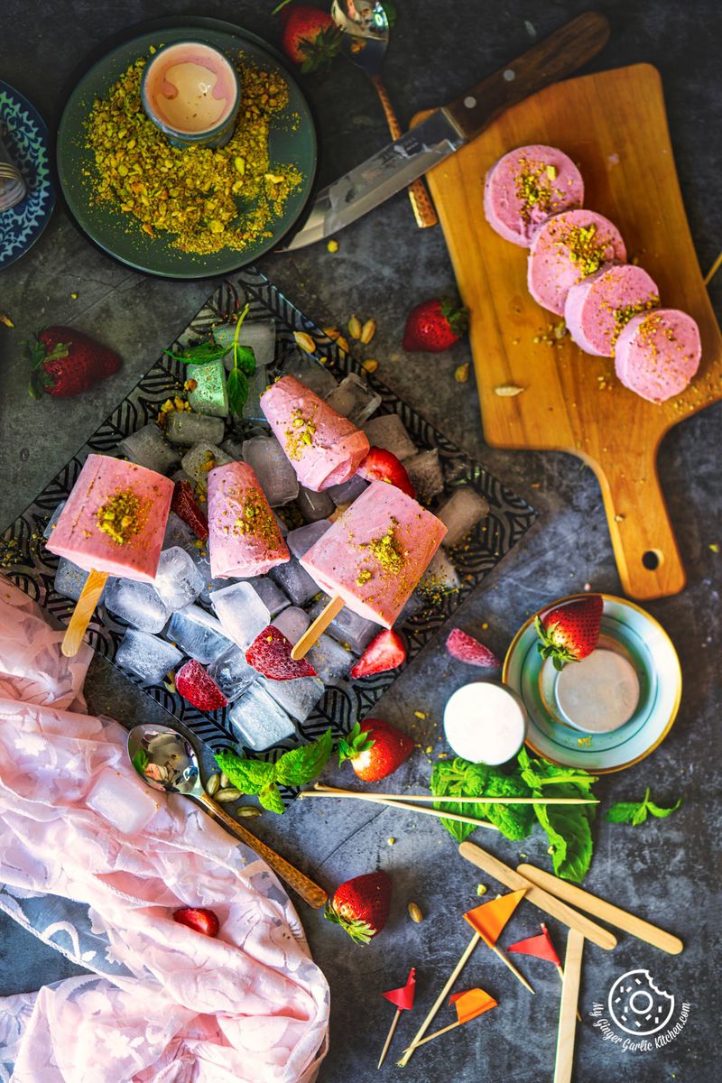 overhead shot of four strawberry kulfis served on a bed of ice cubes, with four roll cut strawberry kulfis on the wooden board