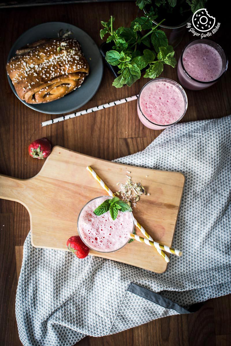 a wooden cutting board with a glass of strawberry lassi with two more strawberry lassi glasses on side