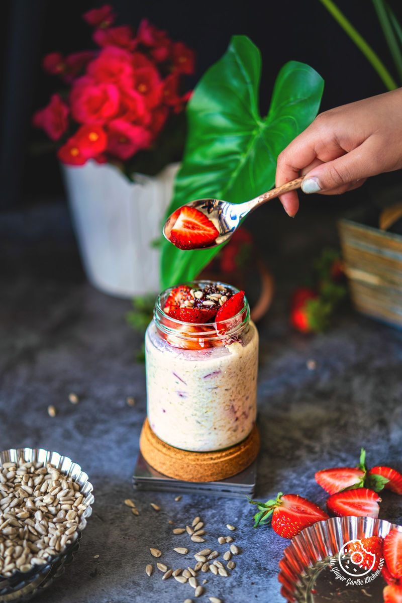 a hand holding a spoon with a sliced strawberry over creamy strawberry overnight oats jar