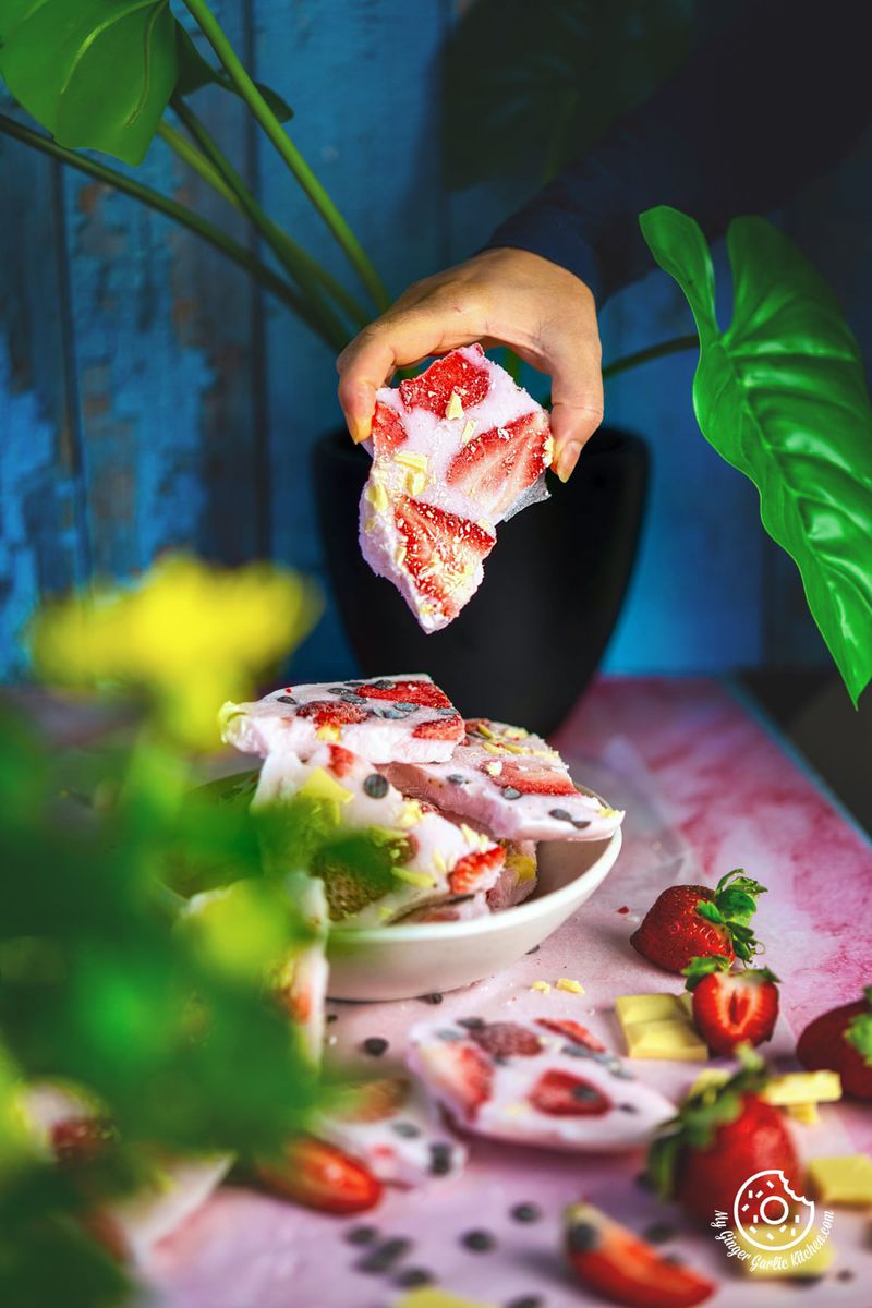 a person holding a piece of strawberry yogurt chocolate bark over a white plate filled with barks