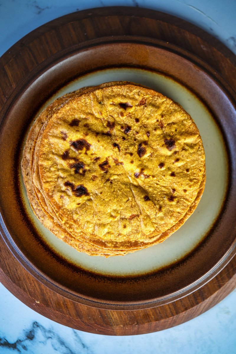 A stack of sweet potato flatbread (roti) is arranged on a brown ceramic plate. The tortillas have a golden orange hue with spots of browning and appear to be cooked evenly.