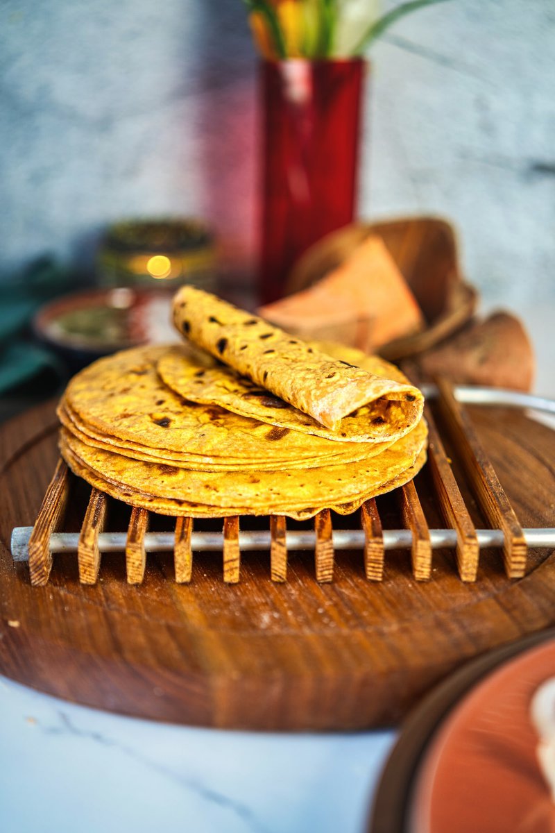 A stack of freshly cooked sweet potato flatbread roti on a wooden rack, rolled for serving, with raw sweet potato in the background.