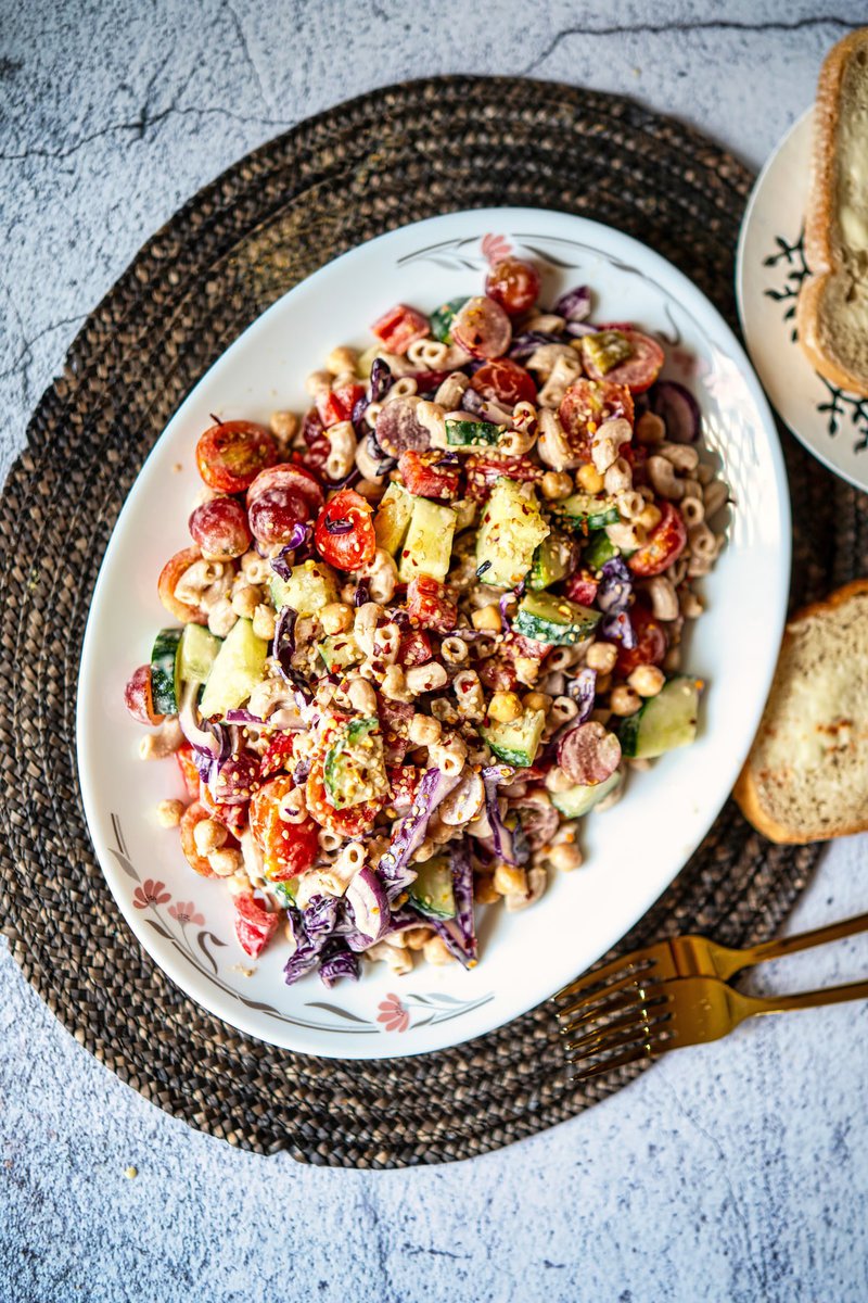 A colorful tahini pasta salad with chopped vegetables on a white oval plate, served on a woven mat.