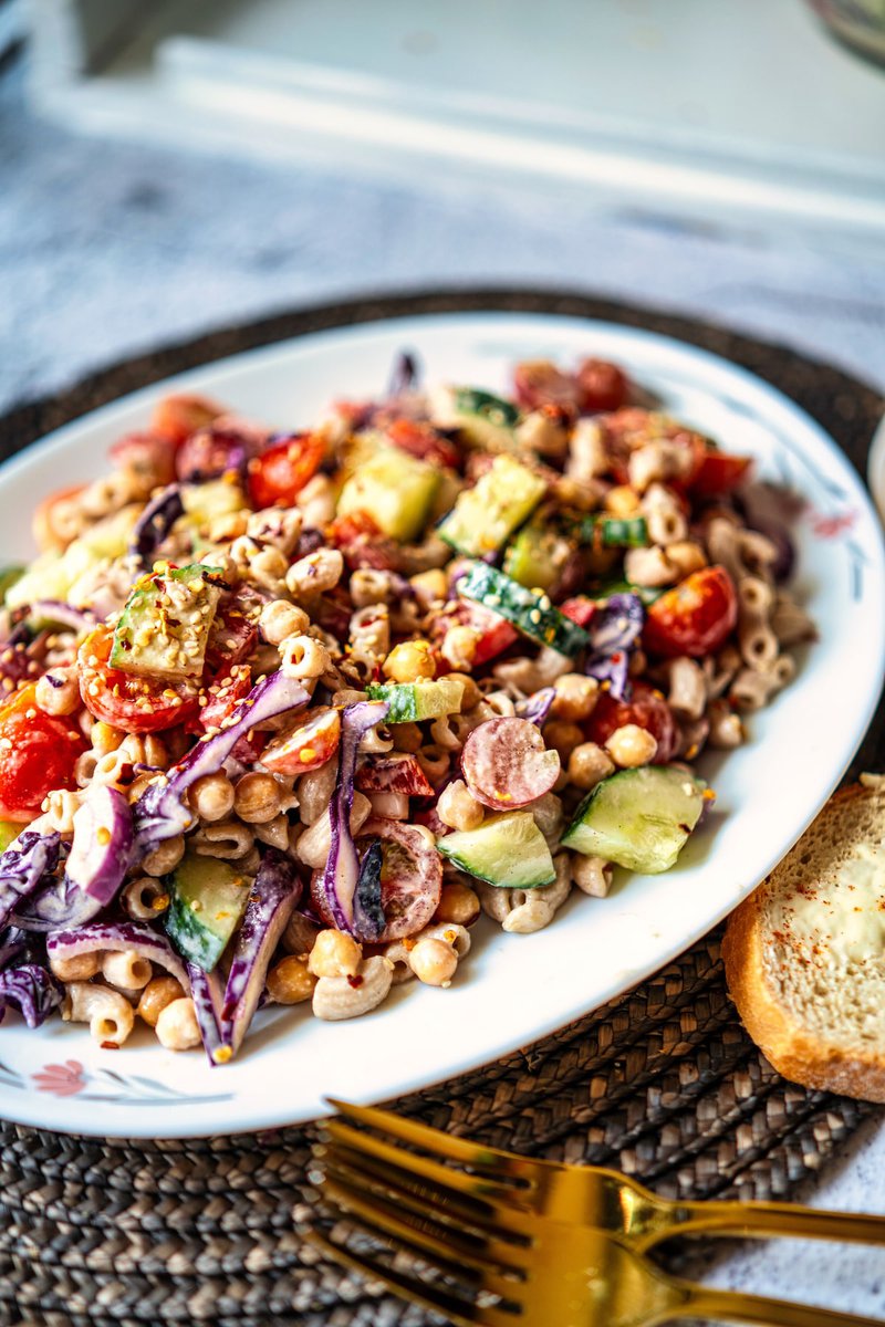 A colorful tahini pasta salad featuring chopped vegetables on a white plate, accompanied by a slice of buttered bread.