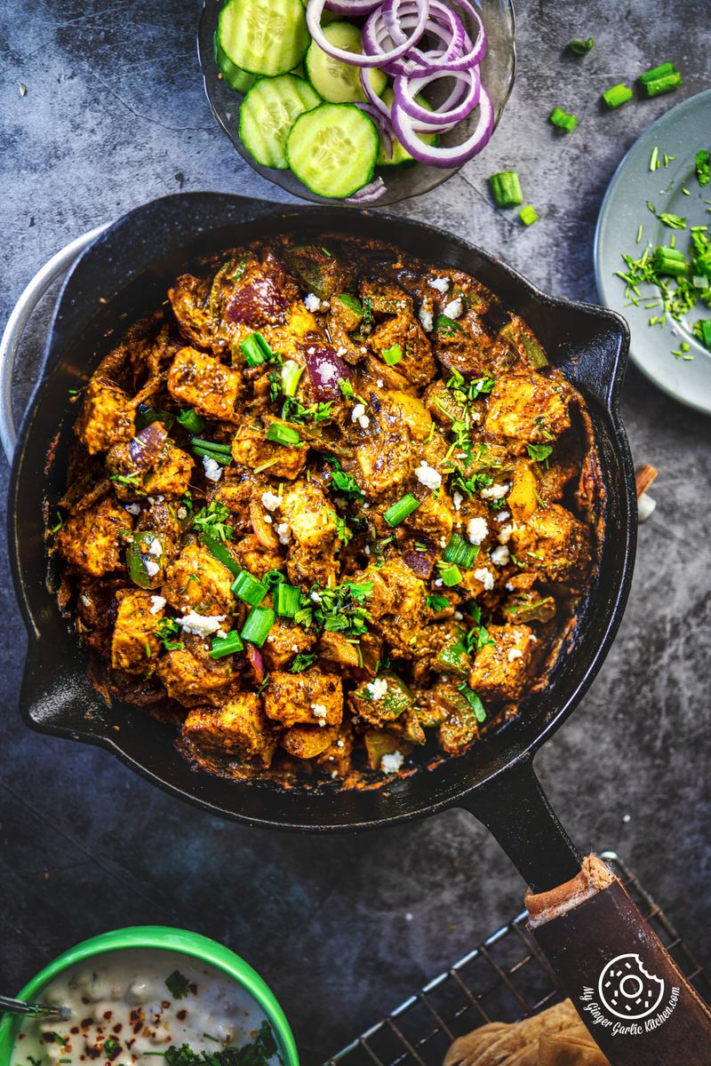 overhead of a cast iron pan of tawa paneer with a spoon next to a bowl of vegetables