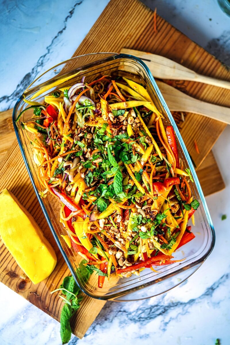 Glass baking dish filled with colorful mango salad, garnished with fresh herbs and nuts, placed on a wooden board.