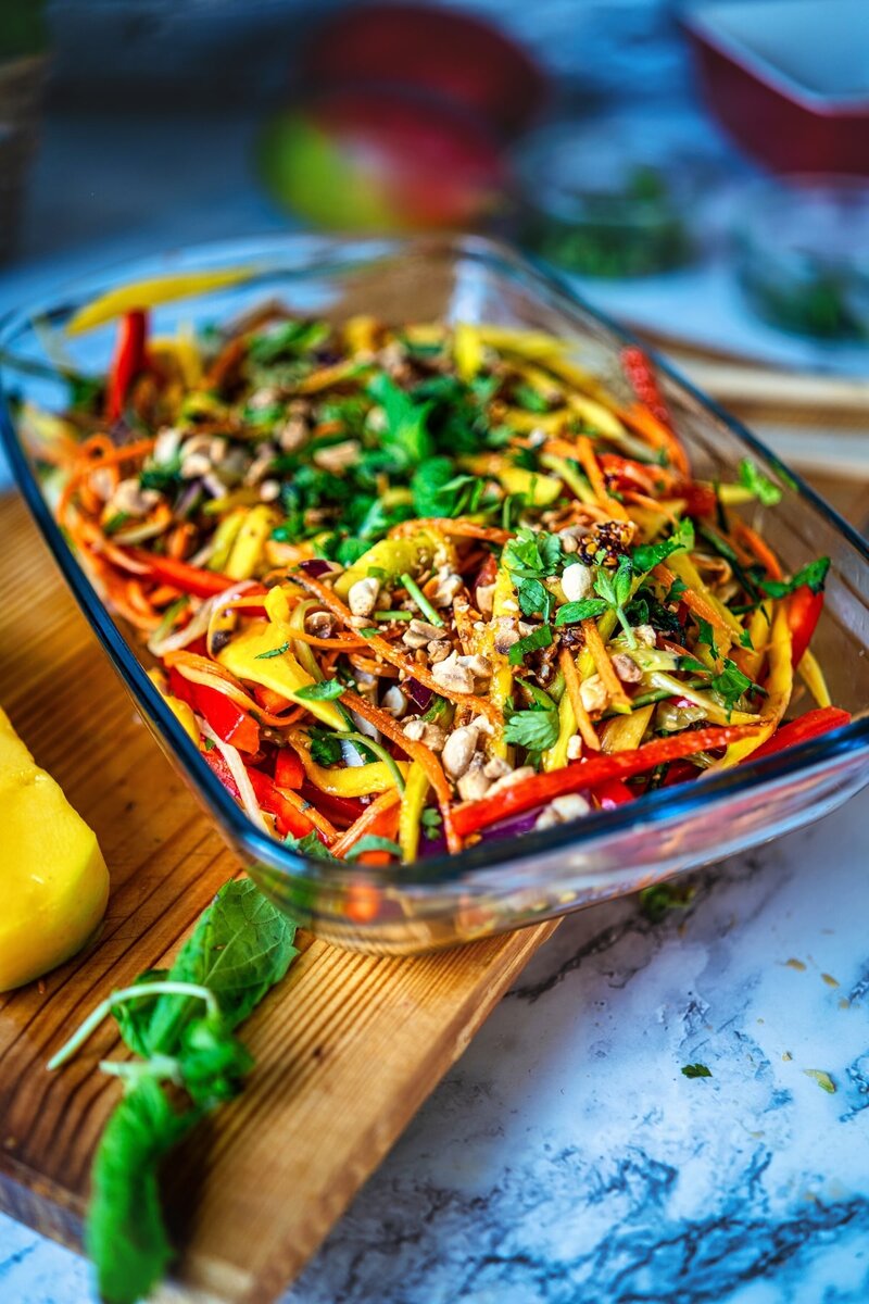 Close-up of fresh mango salad in a glass dish, showing vibrant strips of vegetables and garnished with herbs and nuts.