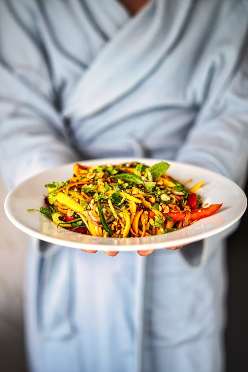 Person in a light robe holding a white plate of fresh mango salad with colorful vegetables and herbs.