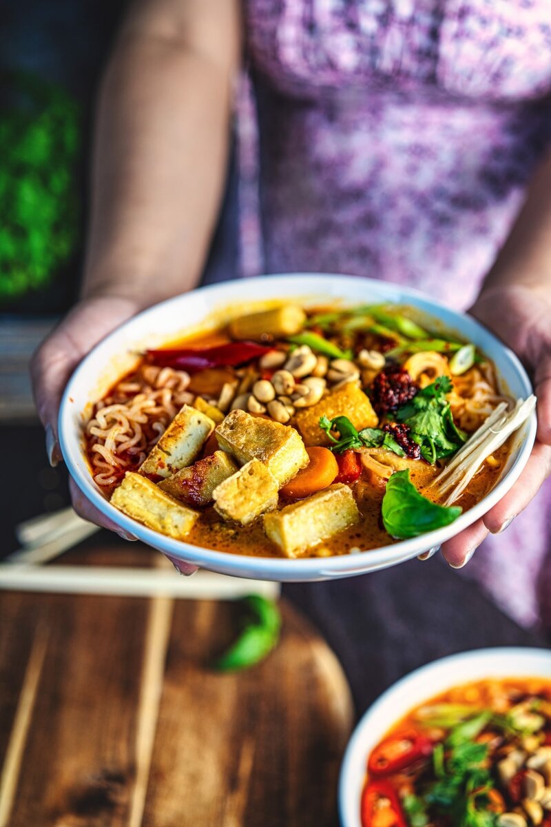 Holding bowl of Thai curry ramen with crispy tofu, vegetables, and creamy coconut red curry broth