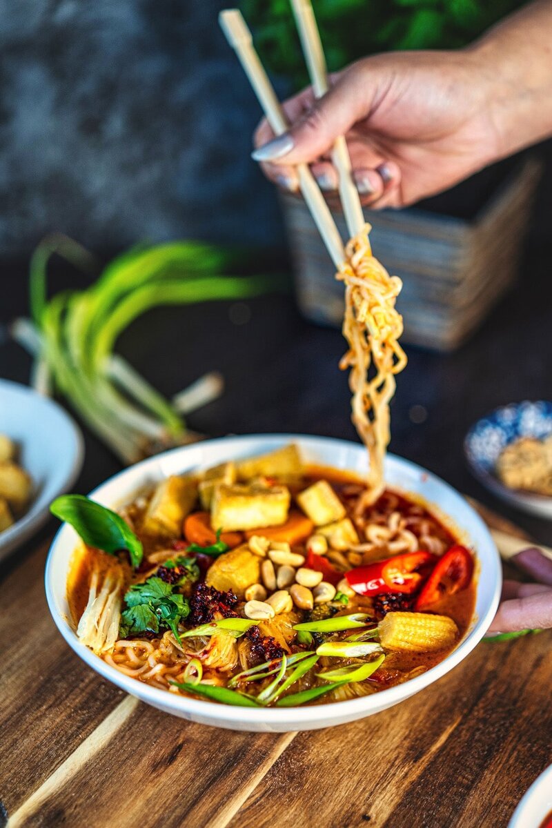 Lifting ramen noodles from a bowl of Thai red curry ramen topped with tofu, peanuts, and fresh herbs
