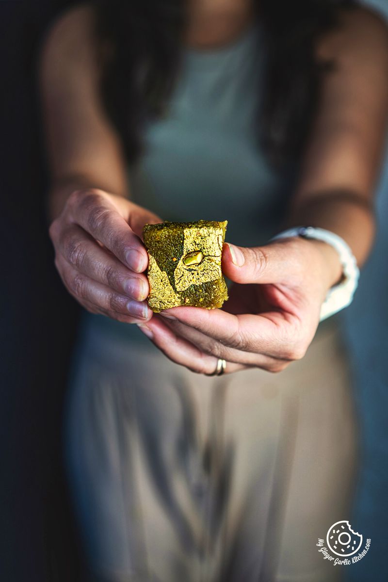 A female holding one piece of Thandai Burfi garnished with edible silver leaves and pistachio