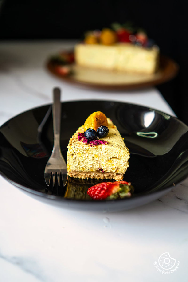 No Bake Thandai Cheesecake slice on a black plate and cheesecake in the background