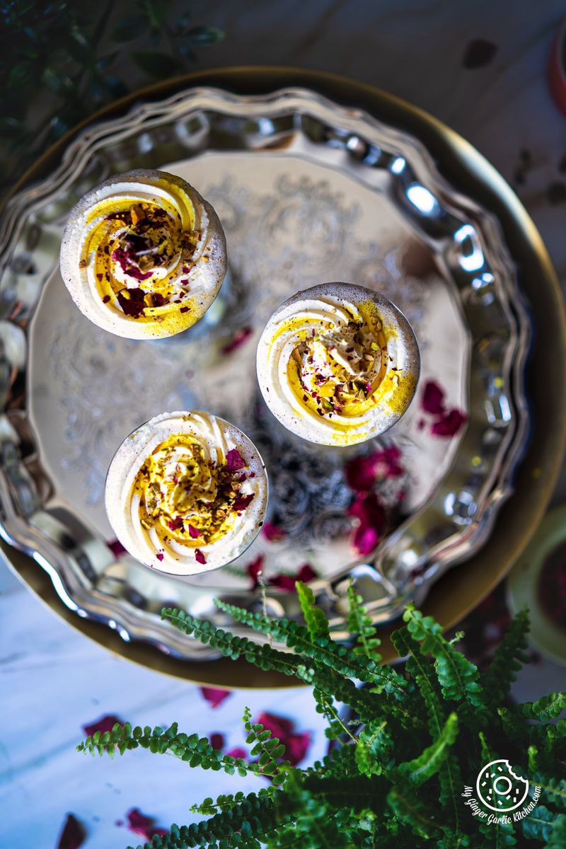 overhead view of 3 glasses of thandai frappe topped with whipped cream and rose petals on a metal plate