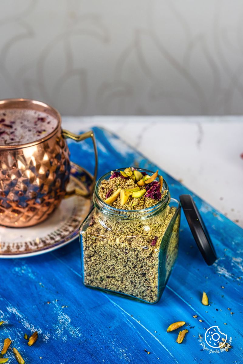 thandai powder in a transparent jar over blue wooden board and a copper mug on the side