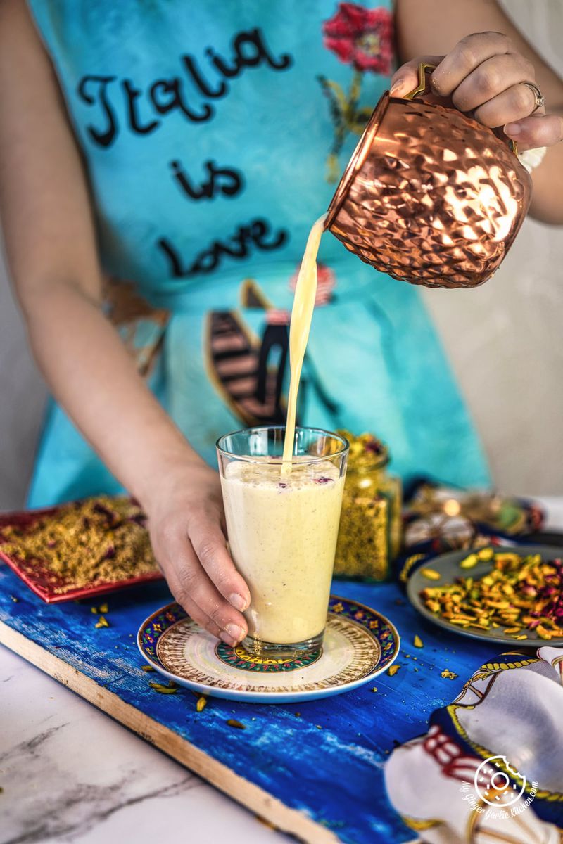 a female pouring thandai in a transparent glass from a copper mug