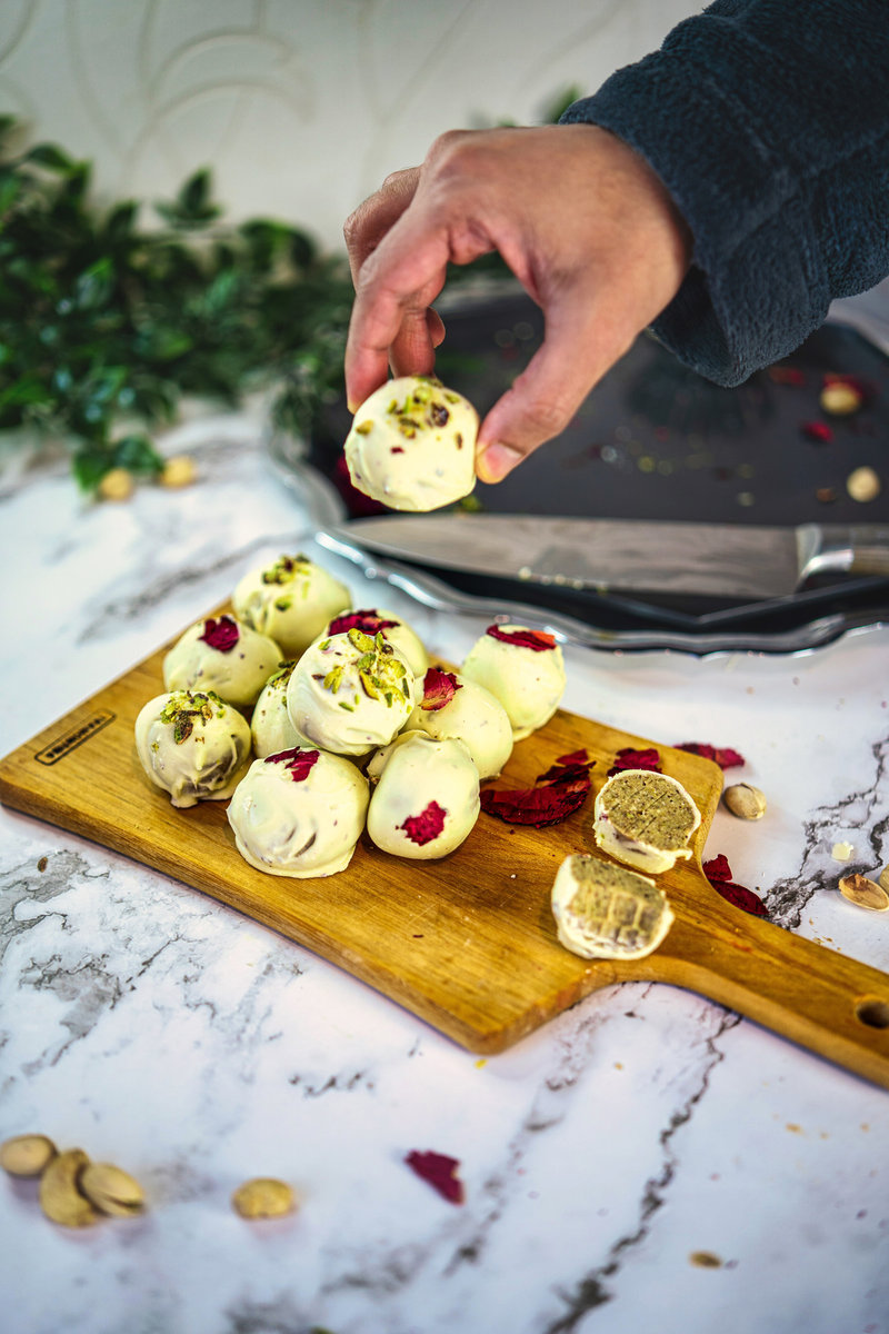 Hand picking a Thandai truffle from a wooden board with a collection of truffles, rose petals, and cardamom pods scattered around.