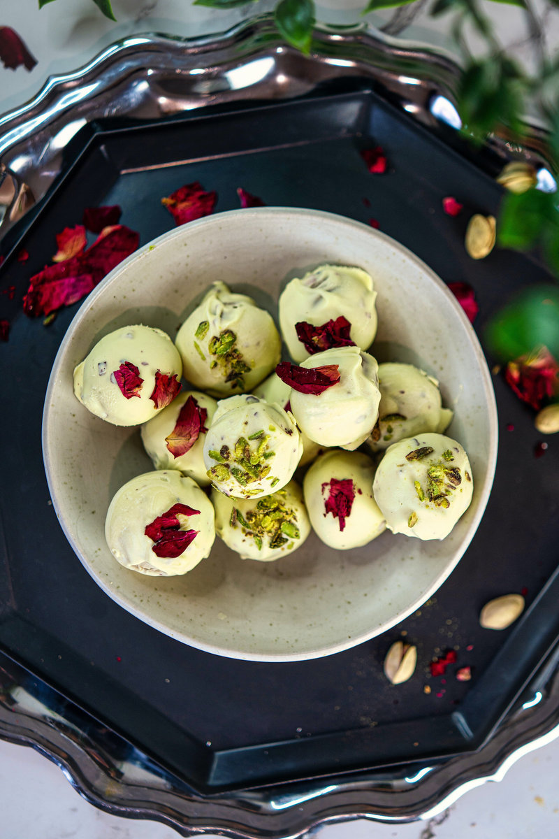 Elegant white bowl filled with Thandai truffles garnished with pistachio and rose petals on a silver tray with scattered ingredients.