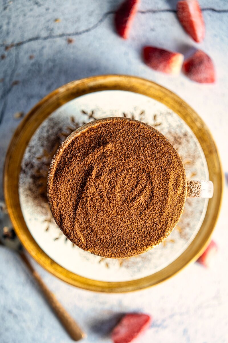 Overhead view of a glass cup filled with tiramisu overnight oats, topped with a layer of cocoa powder, placed on a decorative plate with sliced strawberries in the background.