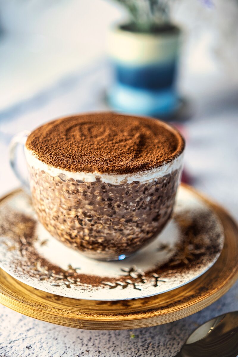 Side view of a cup of tiramisu overnight oats topped with cocoa powder, sitting on a decorative saucer, with a blurred background.