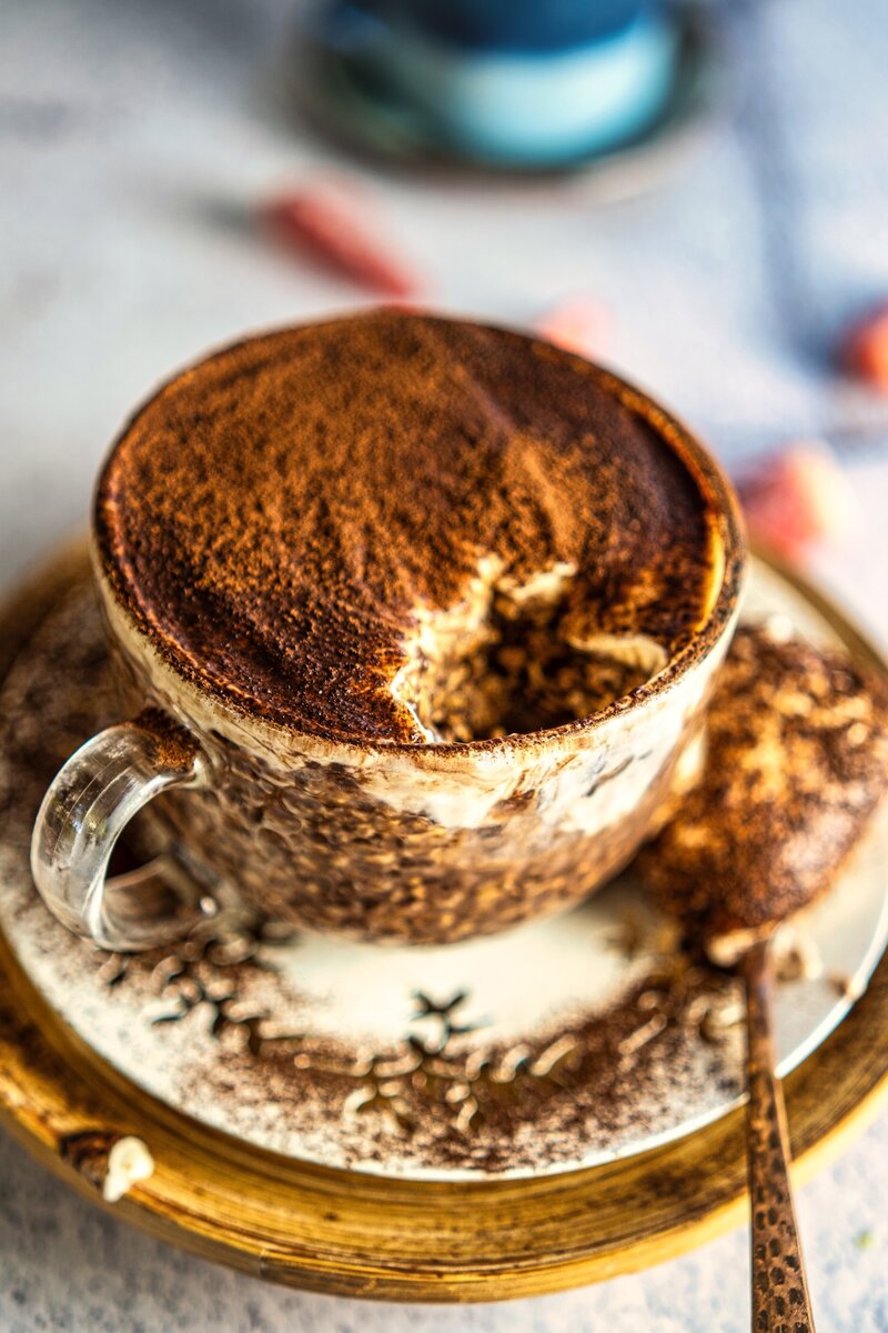 Glass cup of tiramisu overnight oats with a spoonful taken out, showing creamy layers, on a decorative saucer with cocoa powder sprinkled around.