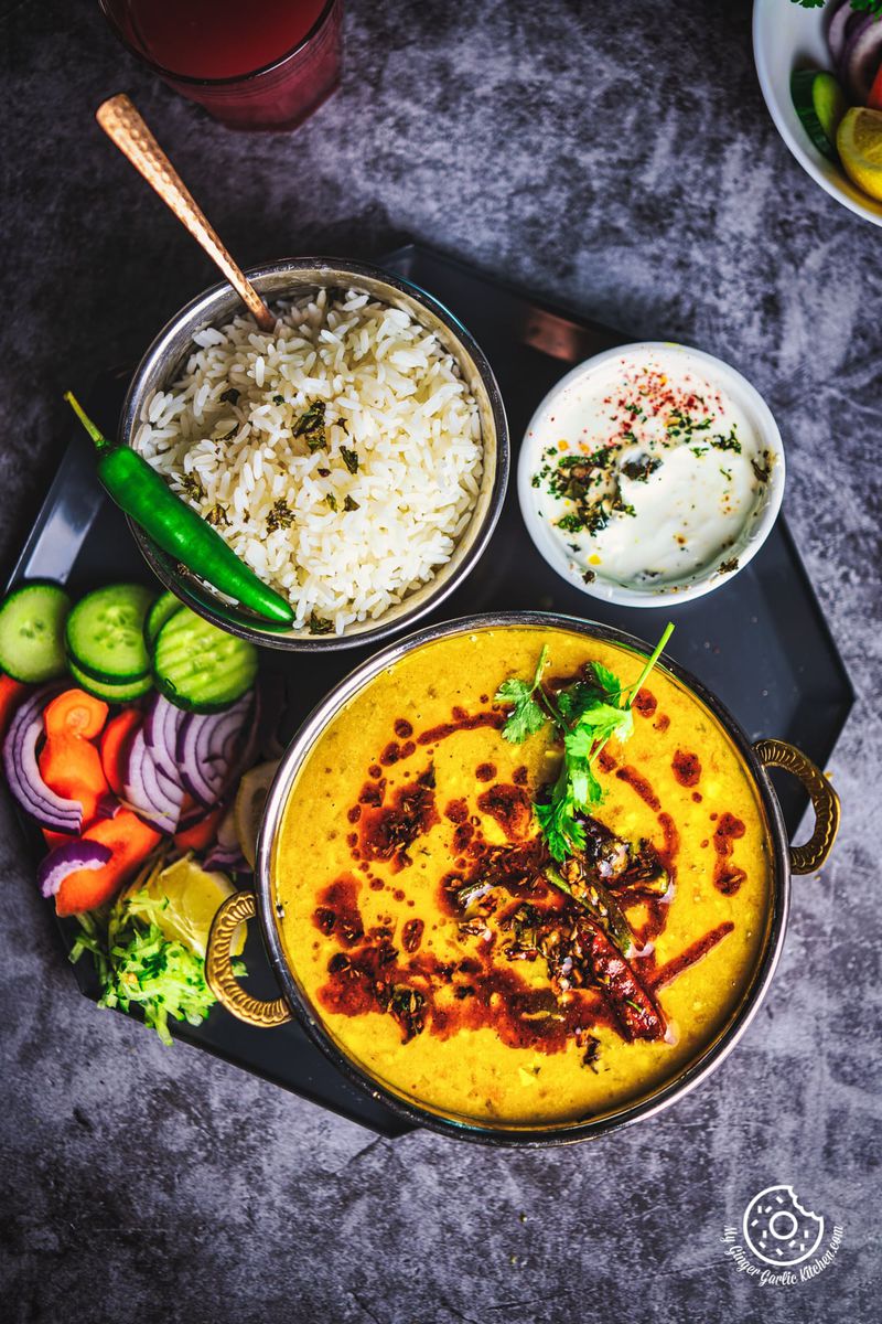 A vibrant presentation of Toor Dal Tadka, with sides of steamed rice, salad, and a dollop of yogurt on a dark textured background.