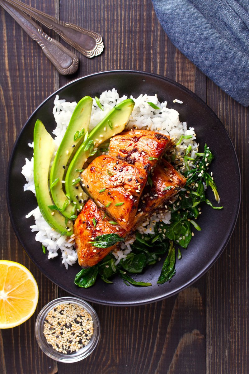 Grilled steak with a side of rice, sliced avocado, and sautéed greens on a black plate, accompanied by a lemon wedge and a small dish of spices.